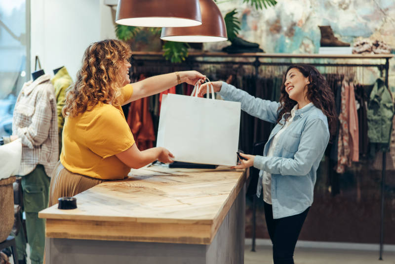 Click and Collect: Eine Verkäuferin übergibt einer Frau Ihren Einkauf im Modegeschäft