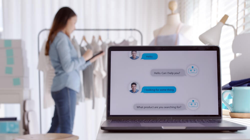 Eine junge Frau verkauft online Mode, auf einem Laptop-Screen unterstützt ein Chatbot einen Kunden bei der Suche nach einem Artikel.