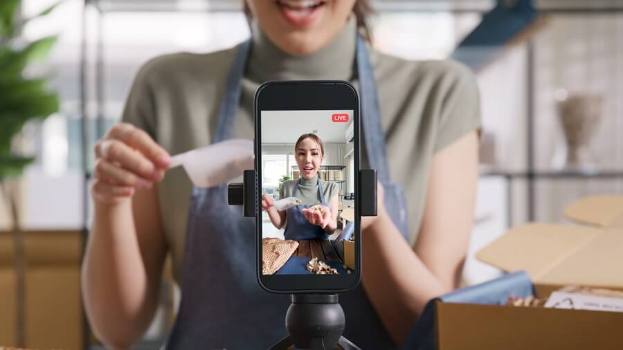 Junge Frau mit blauer Schürze präsentiert Produkte aus ihrem Onlineshop via Smartphone-Kamera im Livestream auf Social Media.