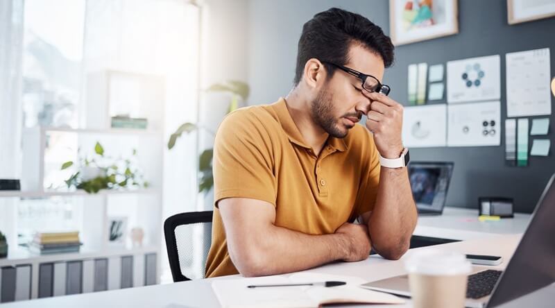 Mann mit Brille und orangefarbenem Polohemd sitzt am Schreibtisch vor einem Laptop und ist ermüdet wegen langen Ladezeiten und schlechter Performance auf Internetseiten