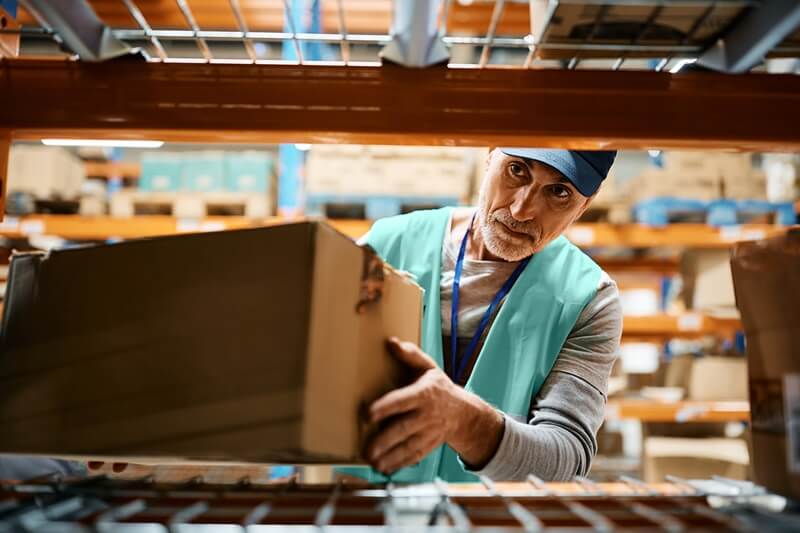 Lagermitarbeiter mit Bart und blauer Schirmmütze stellt ein Paket an den Lagerplatz im Regal