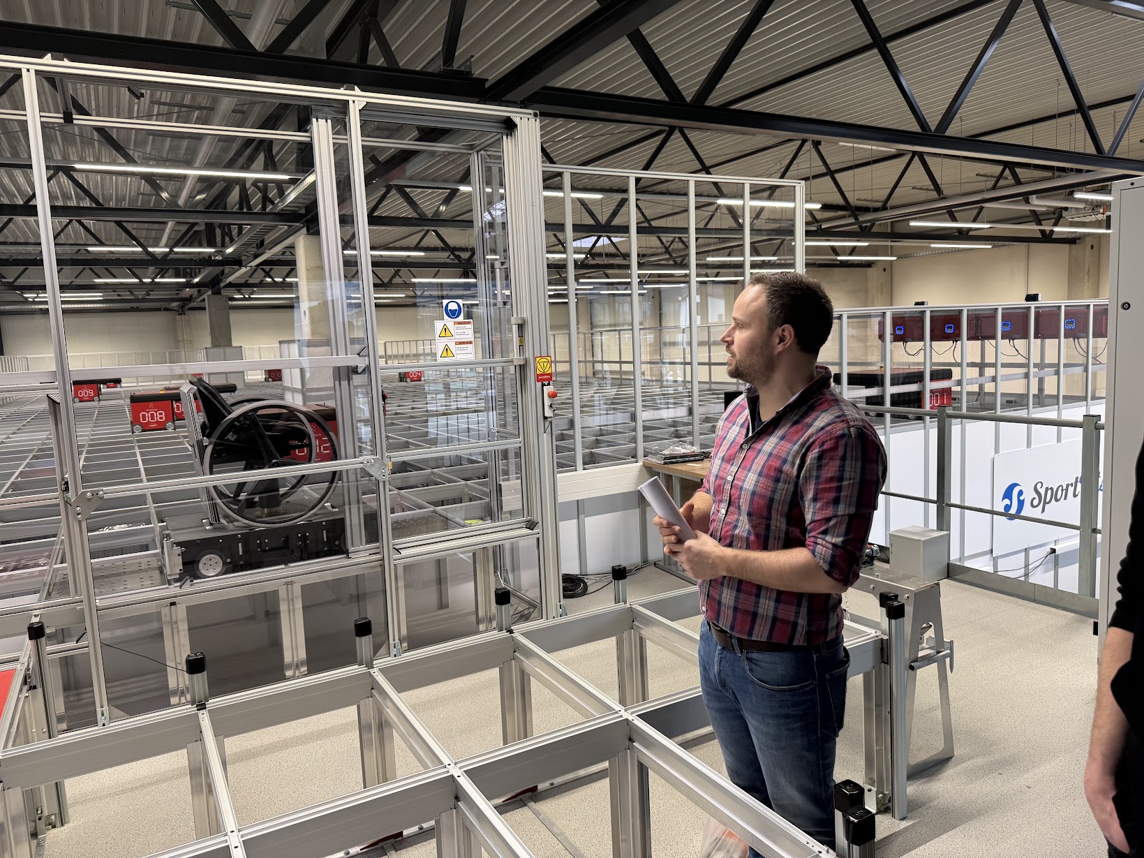 Ein Mann in kariertem Hemd haelt Dokumente und betrachtet ein automatisiertes Lager. Rote Displays zeigen Zahlen, die auf ein Sortiersystem hinweisen.