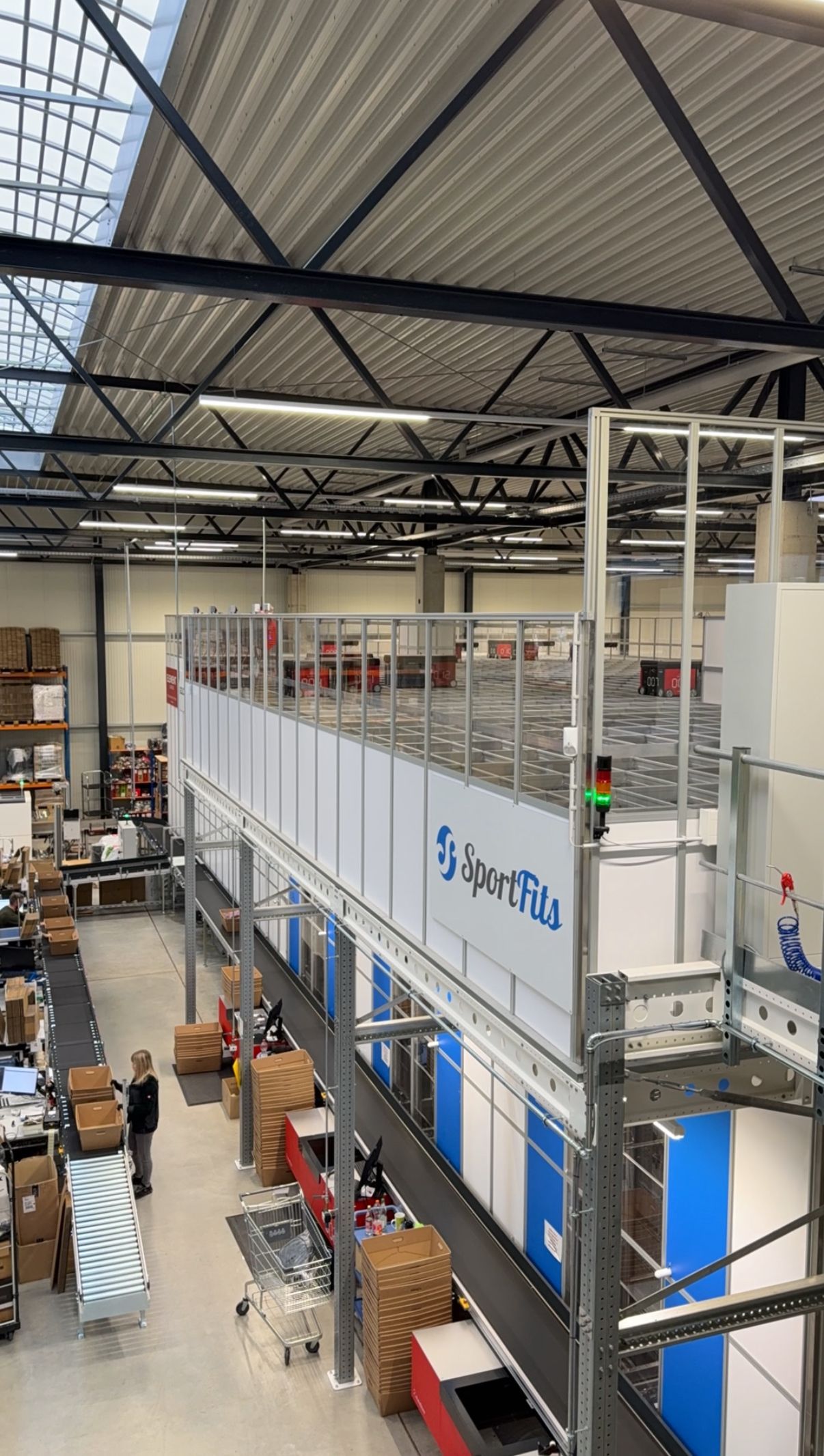 Mehrstoeckiges Lager mit einem AutoStore von „SportFits“. Unten packen Mitarbeitende Waren in Kartons.