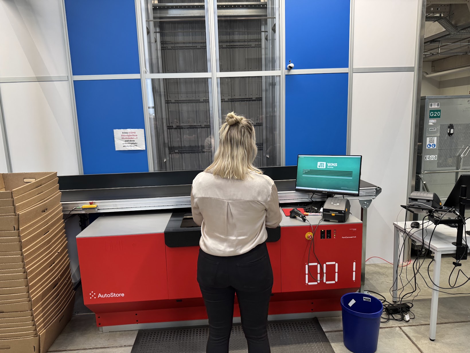 Eine Frau steht vor einem AutoStore-Modul mit roter Front, bedient einen Computer mit „JTL WMS“-Software und scannt Artikel. Kartons und Verpackungsmaterial sind in der Umgebung sichtbar.
