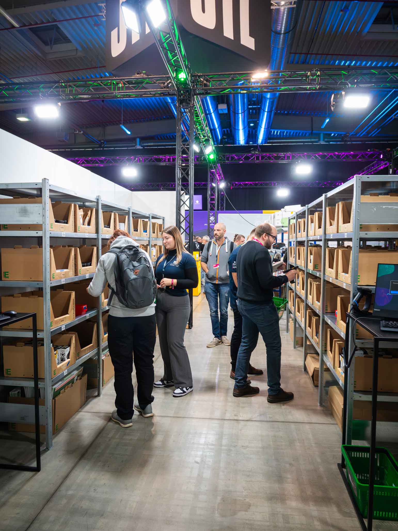 Menschen testen JTL-WMS in einem Regalgang auf einer Messe. Sie inspizieren Produkte in offenen Kartons und nutzen Scanner. Ein „JTL“-Schild haengt über dem Gang, bunte Beleuchtung schafft Messeatmosphaere.
