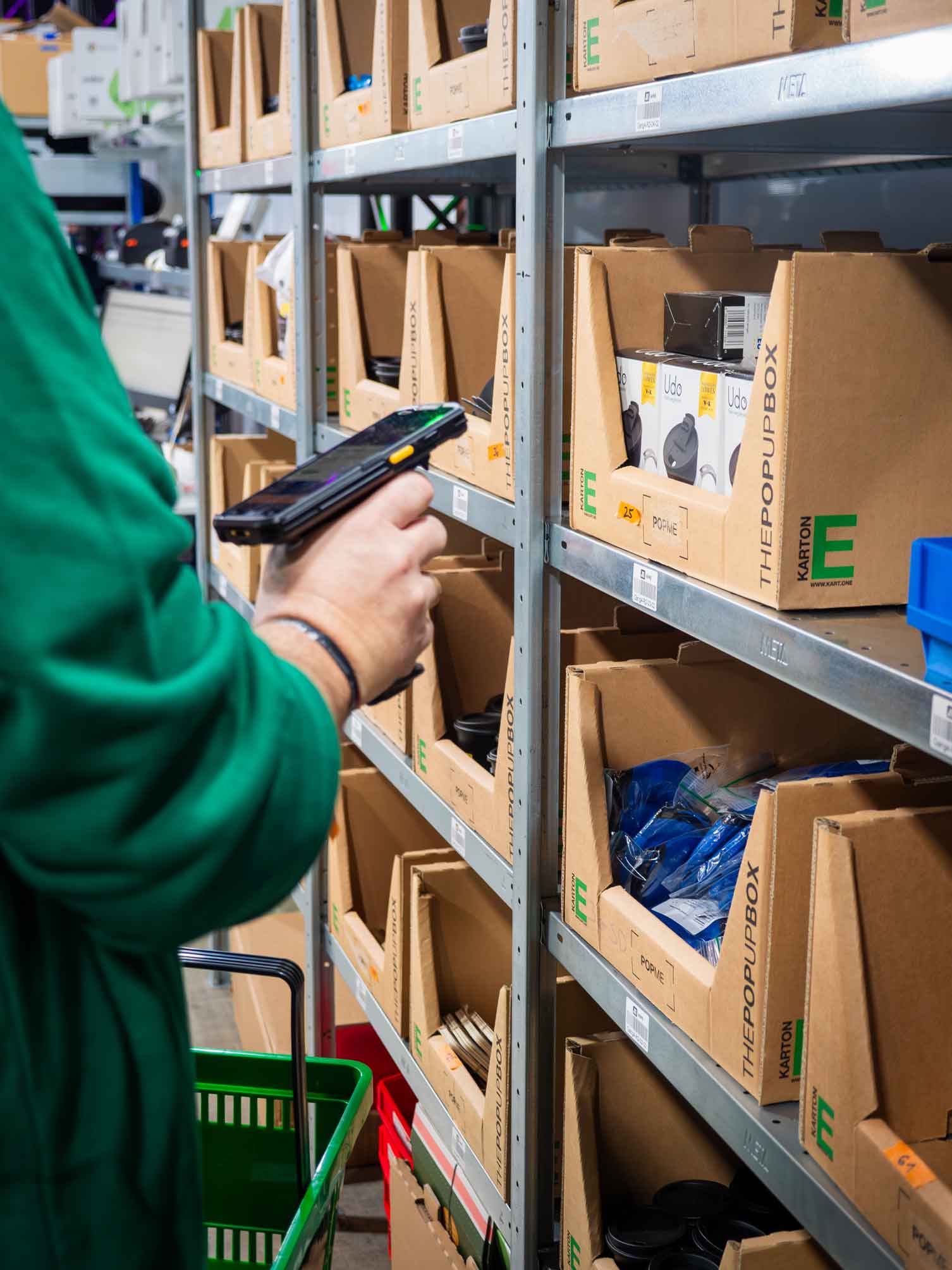 Eine Hand haelt einen Scanner und scannt ein Produkt aus einem offenen Kartonregal mit „THEPOPUPBOX“-Aufdruck. Plastikverpackungen und schwarze Behaelter sind in den Faechern sichtbar.