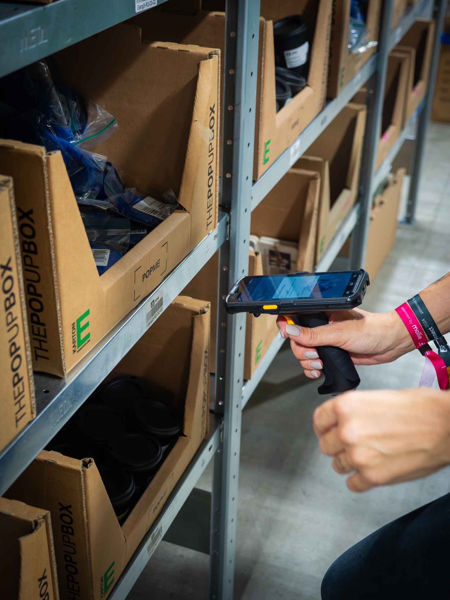 Eine Person scannt Waren in Kartons mit einem Handscanner. Armbaender an ihrem Handgelenk deuten auf eine Messe oder ein Event hin. Das Regal ist mit „THEPOPUPBOX“-Kartonagen bestueckt.