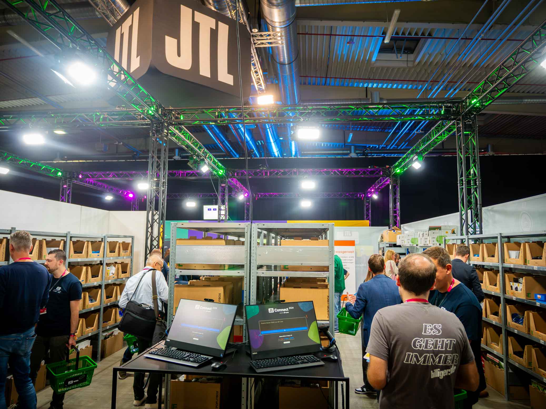 Der JTL-WMS Stand auf der JTL Connect Messe. Besucher mit gruenen Einkaufskoerben testen den Prozess. Bunte Lichter beleuchten die Szene.