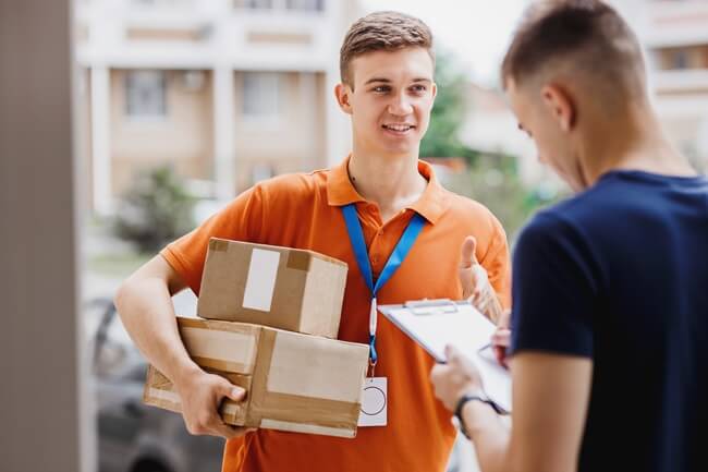 Ein junger Paketzusteller in orangefarbenem Polohemd übergibt zwei Kartons an einen Kunden. Der Zusteller trägt ein Namensschild mit blauem Band und zeigt Daumen hoch. Der Kunde unterschreibt auf einem Klemmbrett. Im Hintergrund sind Wohngebäude unscharf erkennbar.
