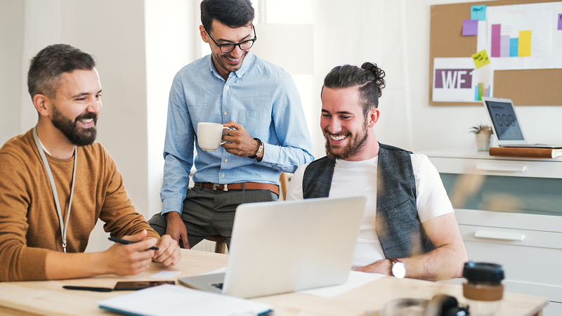 Drei Männer sitzen und stehen um einen Schreibtisch in einem modernen Büro und schauen lachend auf einen Laptop. Auf dem Tisch liegen Notizbücher, Stifte und Kaffeebecher. Im Hintergrund ist eine Pinnwand mit Diagrammen und Post-its zu sehen.
