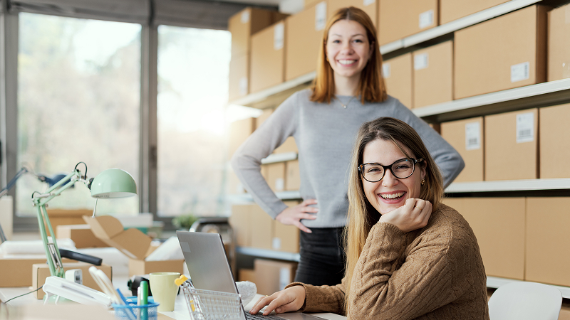 Zwei Frauen befinden sich in einem Raum mit vielen Kartons auf Regalen. Eine sitzt lächelnd an einem Schreibtisch mit Laptop, Büromaterialien und Lampe, die andere steht im Hintergrund und schaut ebenfalls lachend in die Kamera.