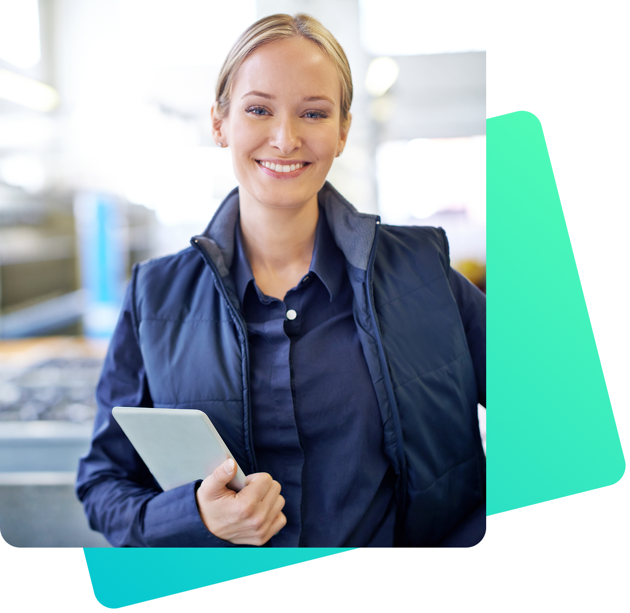 Eine lächelnde, blondhaarige Frau in dunkler Arbeitskleidung und wattierter Weste hält ein Tablet in der Hand. Sie steht in einer hell beleuchteten, industriellen Umgebung mit unscharfem Hintergrund.