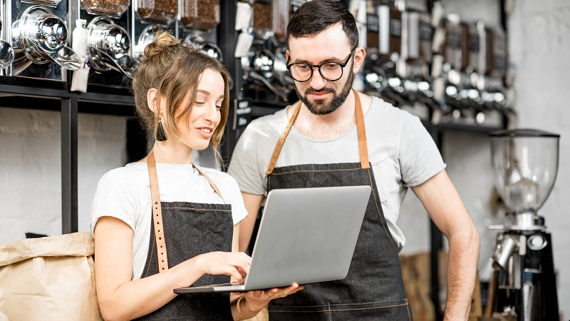 Zwei Personen in Schürzen stehen in einem Laden mit Wandregalen voller Kaffeebohnenbehälter und schauen gemeinsam auf einen Laptop. Die Frau hält das Gerät und tippt, der Mann blickt interessiert auf den Bildschirm.