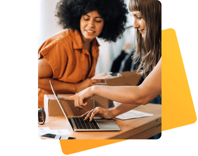 Zwei Frauen arbeiten an einem Tisch mit einem offenen Laptop und Verpackungsmaterial. Eine zeigt auf den Bildschirm, während die andere sich einem offenen Karton zuwendet. Auf dem Tisch liegen Etiketten und ein Etikettendrucker. Die Szene wirkt geschäftig und koordiniert.