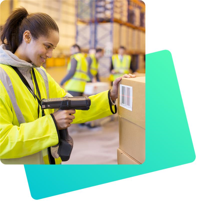 Eine Frau in gelber Warnweste scannt mit einem Handscanner den Barcode eines großen Pakets. Sie steht in einem Lagerhaus, im Hintergrund arbeiten weitere Personen zwischen Regalen und Kartons.