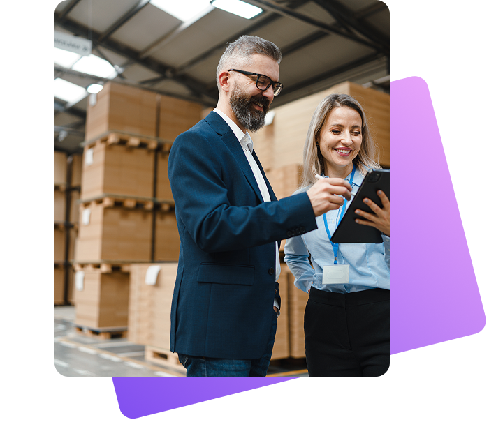 Ein Mann im Anzug und eine Frau in Businesskleidung mit Namensschildern stehen in einem Lager vor gestapelten Paletten und schauen gemeinsam auf ein Tablet. Beide wirken konzentriert und lächeln leicht. Die Halle ist hell und industriell gestaltet.