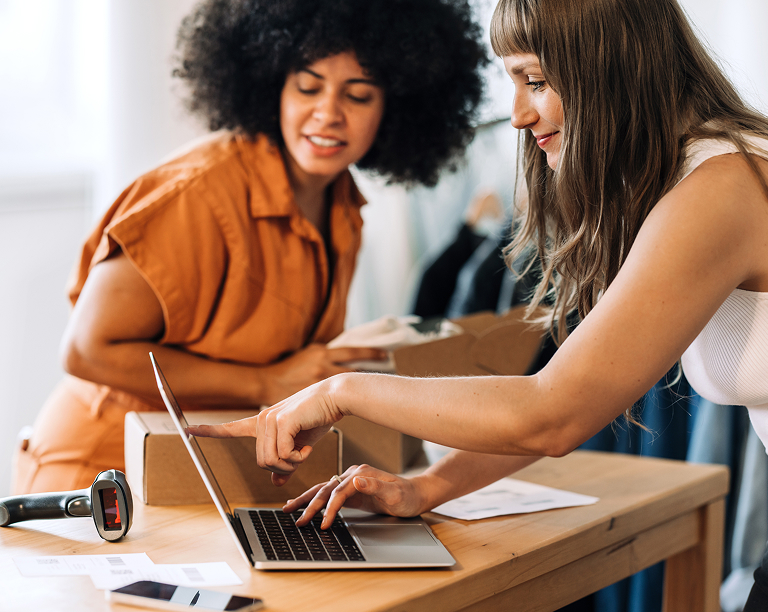 Zwei Frauen arbeiten gemeinsam an einem Tisch mit Laptop, Kartons und Versandetiketten. Eine zeigt auf den Bildschirm, während die andere ein Paket vorbereitet. Auf dem Tisch liegen ein Barcode-Scanner, ein Smartphone und ausgedruckte Etiketten. Im Hintergrund hängen Kleidungsstücke auf einem Kleiderständer.