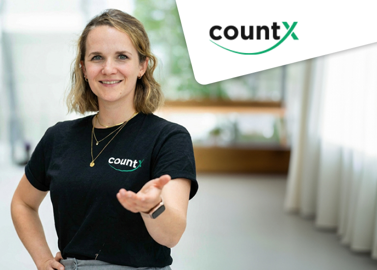 Eine lächelnde Frau steht in einem hellen, modernen Raum und trägt ein schwarzes T-Shirt mit dem Logo „countX“. Sie streckt eine Hand einladend nach vorn aus, als wolle sie jemanden begrüßen oder etwas erklären. In der oberen rechten Ecke ist das countX-Logo zusätzlich eingeblendet.