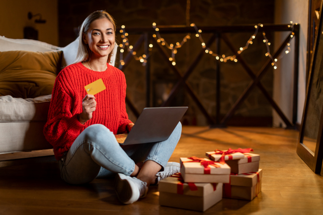 Junge Frau zu Hause mit Weihnachtsbeleuchtung hält Kreditkarte, am Laptop mit Weihnachtspaketen