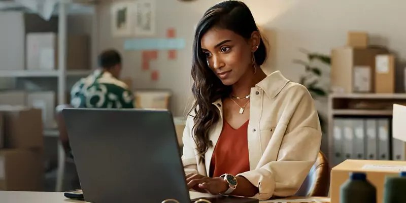 Eine Frau arbeitet an einem Laptop in einem Büro mit Paketen und Ordnern im Hintergrund. Im Hintergrund ist eine weitere Person an einem Arbeitsplatz zu sehen.