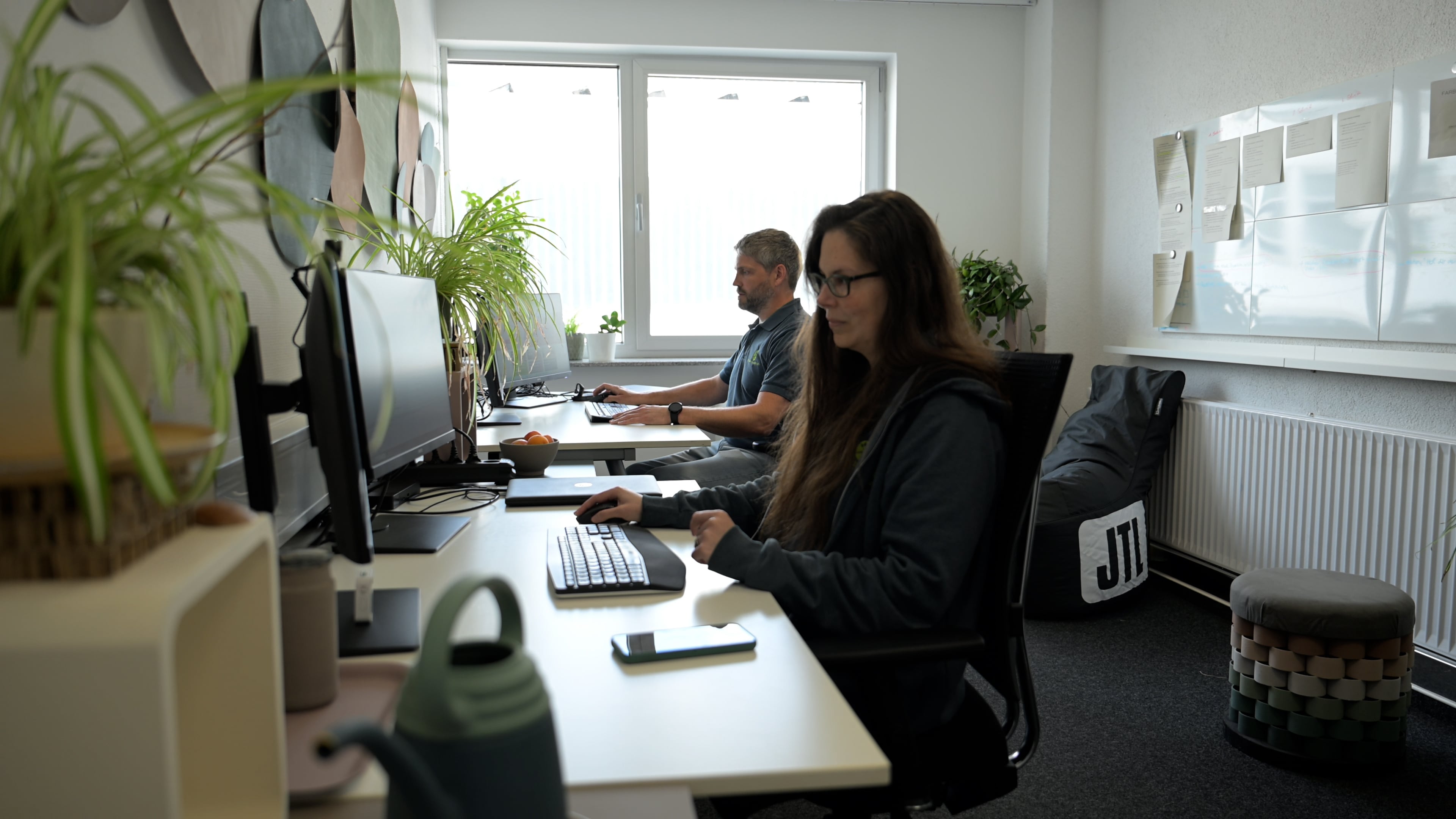Zwei Kolleg:innen arbeiten konzentriert an ihren Schreibtischen im Büro, umgeben von Pflanzen und mit einem JTL-Sitzsack im Hintergrund.